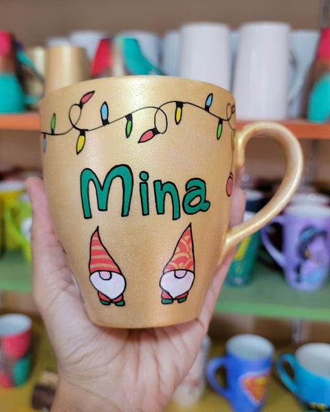 A Gold hand painted mug with gnomes all around the bottom half of the mug and festive lighting all over the top of the mug and the name Mina in the center