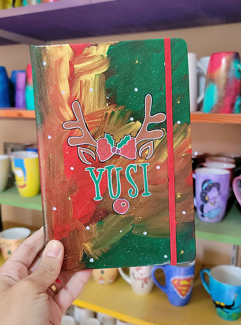 An A5 notebook hand painted in christmas Galaxy background  with reindeer antlers with a bow in the middle and reindeer ears then below them the name "Yusi" and then below it the reindeer nose. all with white dots and gold stars scattered all over