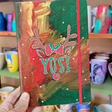 An A5 notebook hand painted in christmas Galaxy background  with reindeer antlers with a bow in the middle and reindeer ears then below them the name 