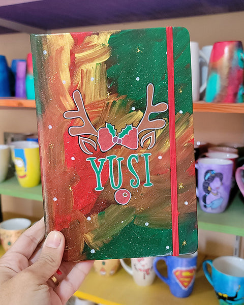 An A5 notebook hand painted in christmas Galaxy background  with reindeer antlers with a bow in the middle and reindeer ears then below them the name "Yusi" and then below it the reindeer nose. all with white dots and gold stars scattered all over