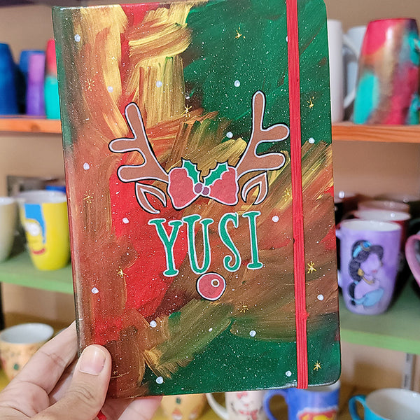 An A5 notebook hand painted in christmas Galaxy background  with reindeer antlers with a bow in the middle and reindeer ears then below them the name "Yusi" and then below it the reindeer nose. all with white dots and gold stars scattered all over