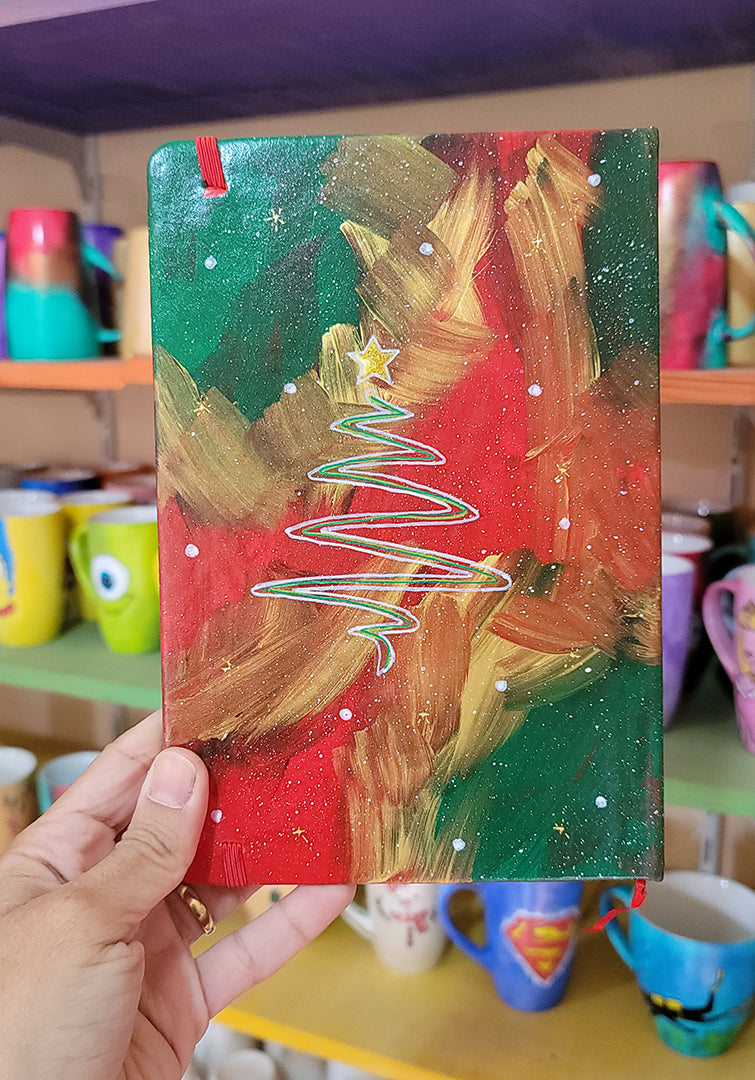 An A5 notebook hand painted in christmas Galaxy background  with a christmas tree drawn in white and colored in green and a gold star on top. all with white dots and gold stars scattered all over