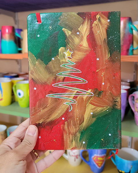 An A5 notebook hand painted in christmas Galaxy background  with a christmas tree drawn in white and colored in green and a gold star on top. all with white dots and gold stars scattered all over