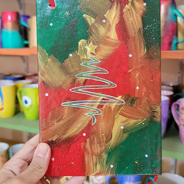 An A5 notebook hand painted in christmas Galaxy background  with a christmas tree drawn in white and colored in green and a gold star on top. all with white dots and gold stars scattered all over