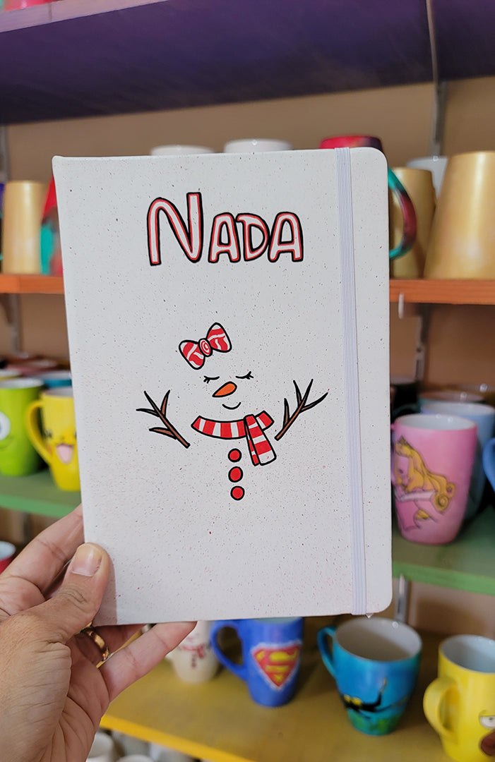 A White A5 notebook with the name nada written on the top center in black and red and a red bow drawn with 2 eyes closed and an orange nose and a smiley mouth with a red scarf and 3 red buttons below it. and tree branches drawn as hands