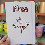 A White A5 notebook with the name nada written on the top center in black and red and a red bow drawn with 2 eyes closed and an orange nose and a smiley mouth with a red scarf and 3 red buttons below it. and tree branches drawn as hands