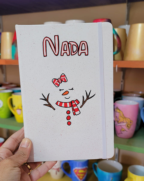 A White A5 notebook with the name nada written on the top center in black and red and a red bow drawn with 2 eyes closed and an orange nose and a smiley mouth with a red scarf and 3 red buttons below it. and tree branches drawn as hands