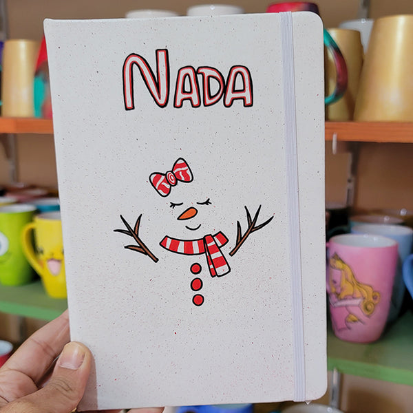 A White A5 notebook with the name nada written on the top center in black and red and a red bow drawn with 2 eyes closed and an orange nose and a smiley mouth with a red scarf and 3 red buttons below it. and tree branches drawn as hands