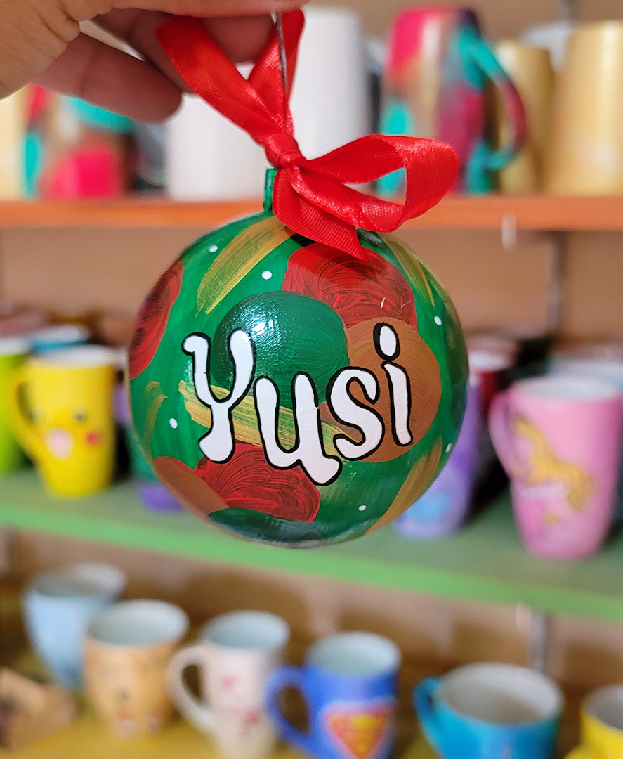 A Colorful hand painted ornament with green background and colors of red green, brown and gold scattered all over with white dots and the name "Yusi" written in white outlined with black