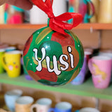 A Colorful hand painted ornament with green background and colors of red green, brown and gold scattered all over with white dots and the name 