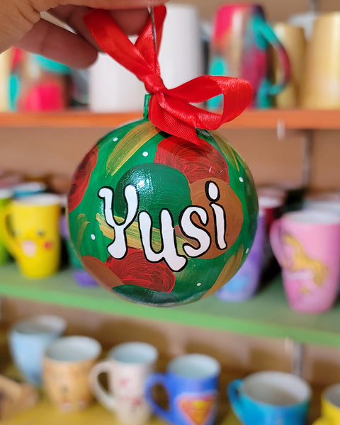 A Colorful hand painted ornament with green background and colors of red green, brown and gold scattered all over with white dots and the name "Yusi" written in white outlined with black