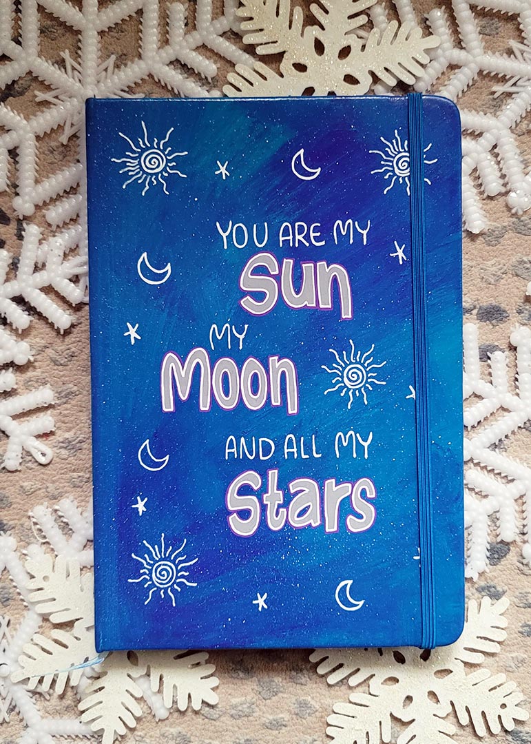 A Blue Galaxy Hand painted Notebook with the sentence "you are my Sun, My Moon and all my stars" with small moons stars and suns scattered all over the mug