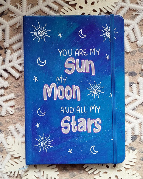 A Blue Galaxy Hand painted Notebook with the sentence "you are my Sun, My Moon and all my stars" with small moons stars and suns scattered all over the mug