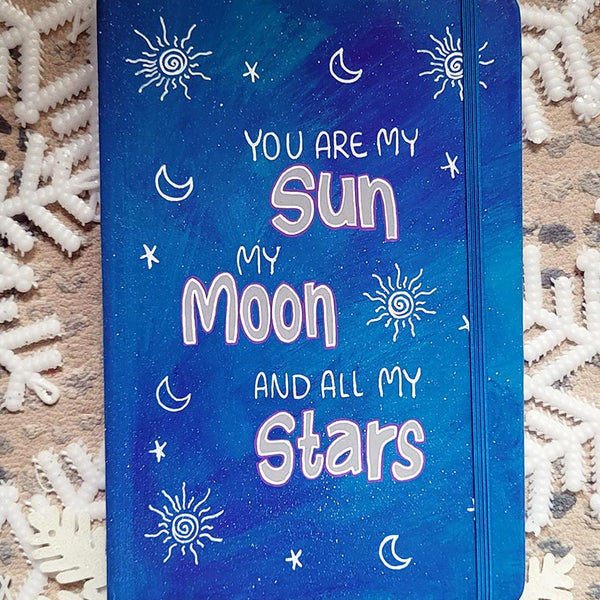 A Blue Galaxy Hand painted Notebook with the sentence "you are my Sun, My Moon and all my stars" with small moons stars and suns scattered all over the mug