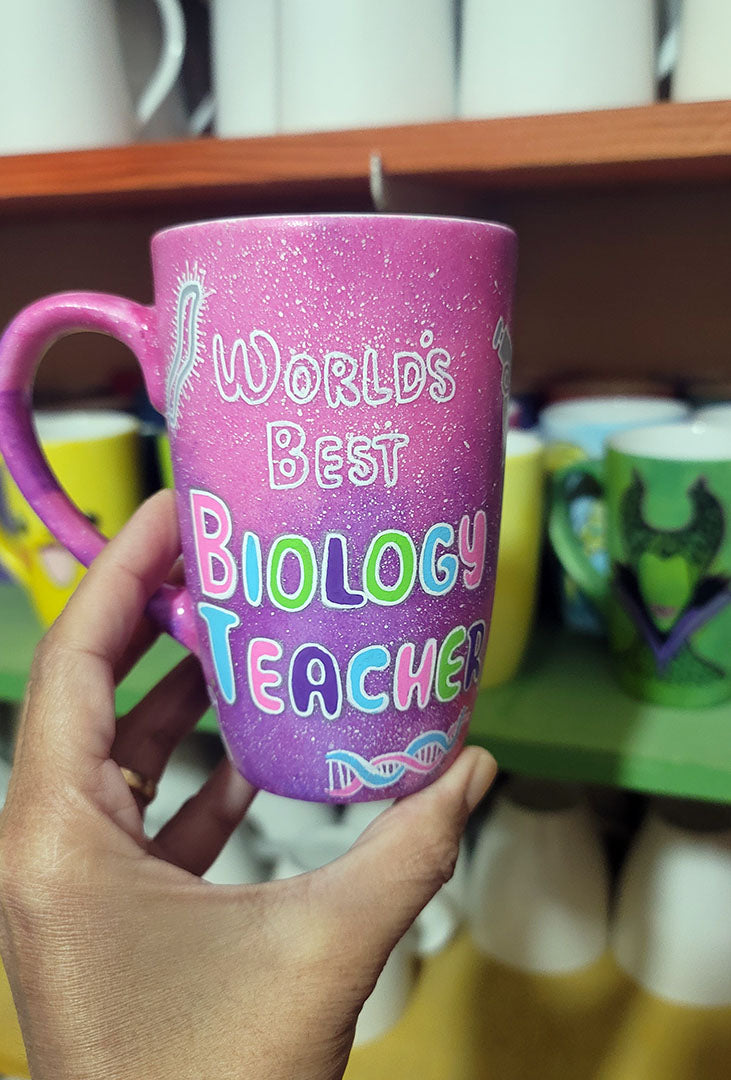 A Pink Galaxy hand painted mug with the words " worlds best biology teacher" and biology items scattered all over (DNA, Plant cell, microscope...etc.)