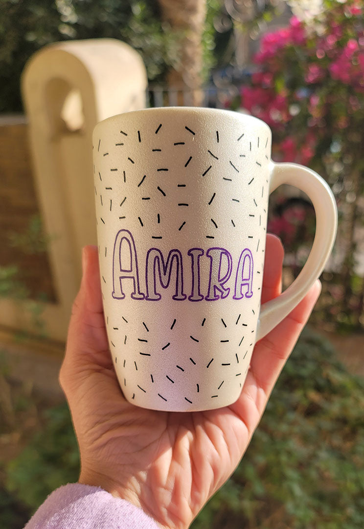 A white pearl hand painted mug with the name "Amira " in purple and black sprinkles scattered all over the mug