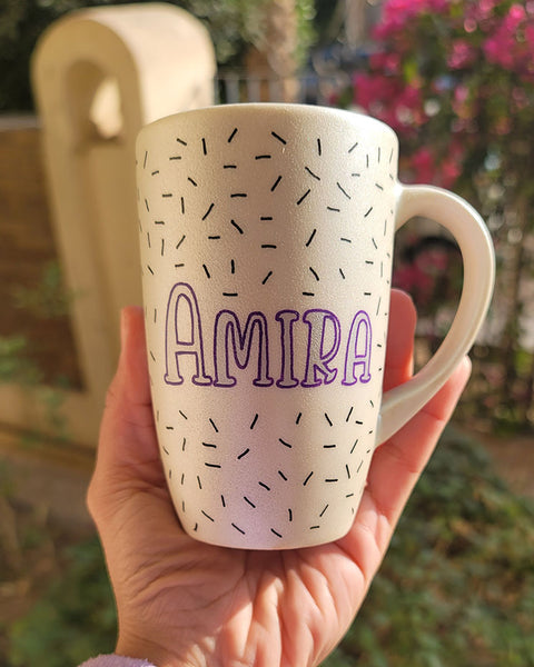 A white pearl hand painted mug with the name "Amira " in purple and black sprinkles scattered all over the mug