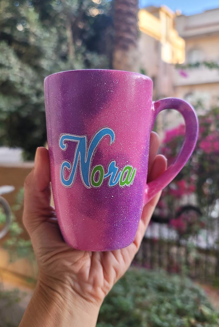 A hand painted pink galaxy mug with the name "Nora"