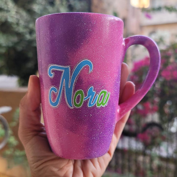 A hand painted pink galaxy mug with the name "Nora"