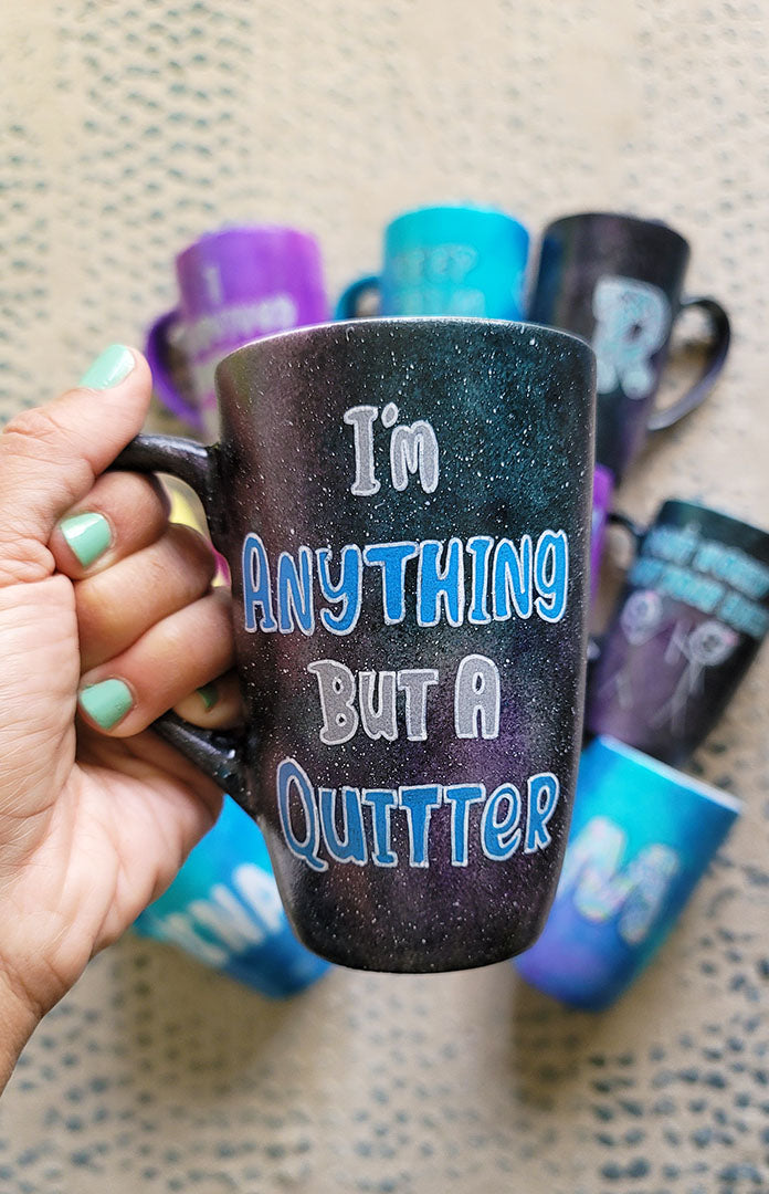 A Black galaxy hand painted mug with the sentence "I'm Anything but a quitter"