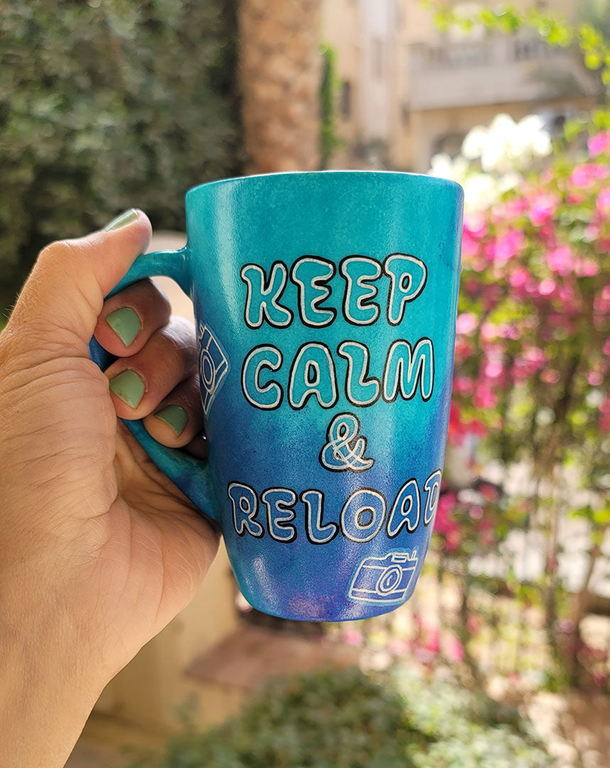 A Blue hand painted mug with the words "Keep Calm and reload" and small cameras scattered all over the mug