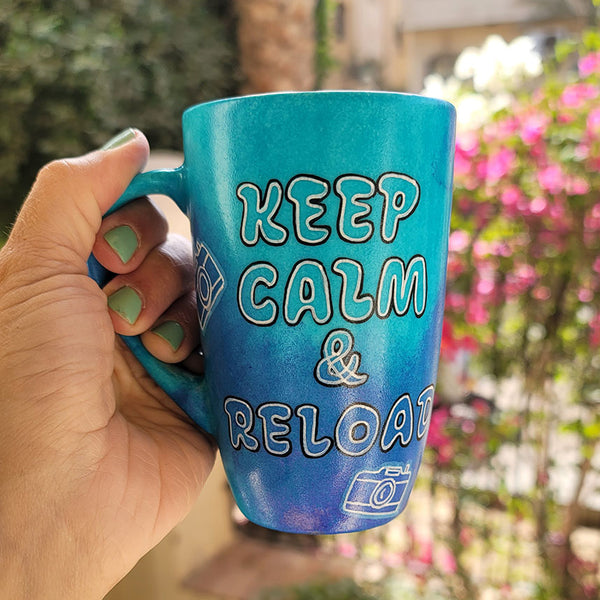 A Blue hand painted mug with the words "Keep Calm and reload" and small cameras scattered all over the mug