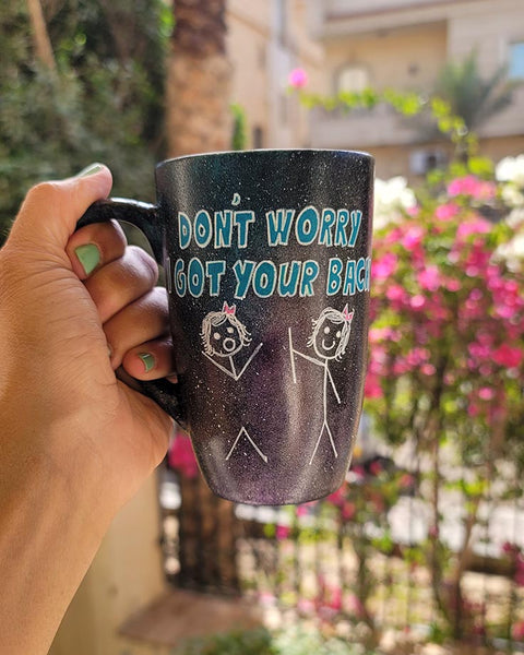 A hand painted black galaxy mug with the words "don't worry, I got your back " and a drawing of 2 stick figures with the girl holding the back of the other girl