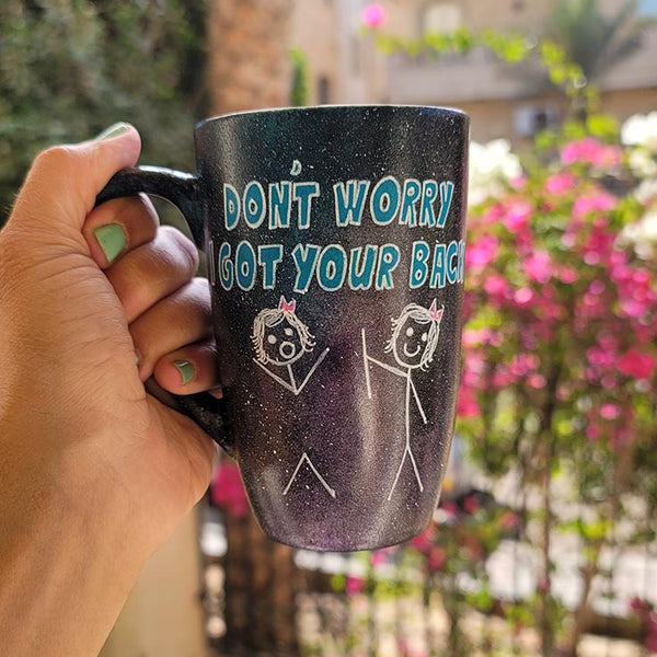 A hand painted black galaxy mug with the words "don't worry, I got your back " and a drawing of 2 stick figures with the girl holding the back of the other girl