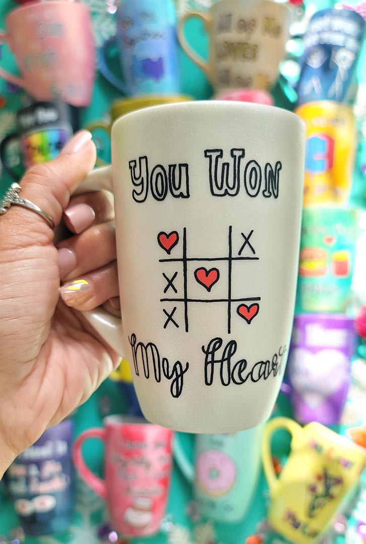 A hand painted pearl white mug with the sentence "you won" and under it an x o chart with hearts instead of o  and three hearts winning the game diagonally, then under it the words "My heart"