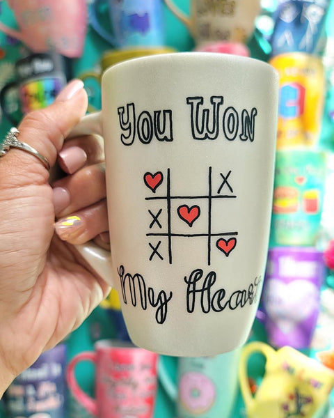 A hand painted pearl white mug with the sentence "you won" and under it an x o chart with hearts instead of o  and three hearts winning the game diagonally, then under it the words "My heart"