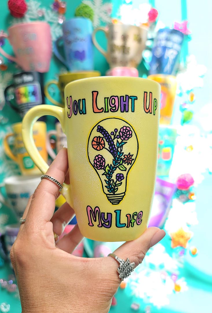 A Pastel yellow hand-painted mug with the words "you light up my life" and a bulb drawn on it with flowers inside