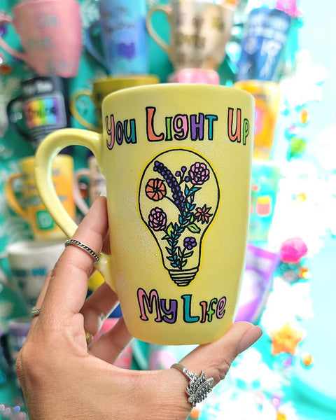 A Pastel yellow hand-painted mug with the words "you light up my life" and a bulb drawn on it with flowers inside