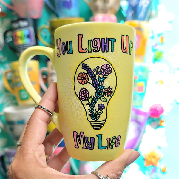 A Pastel yellow hand-painted mug with the words "you light up my life" and a bulb drawn on it with flowers inside