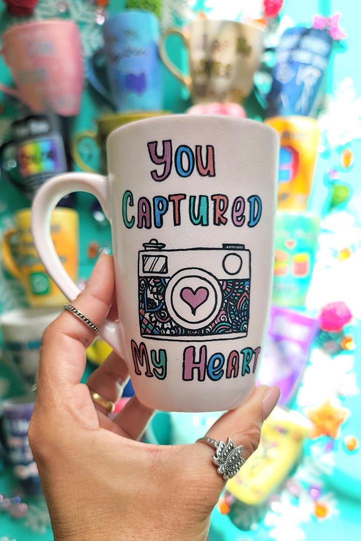 A pale pink mug with the words "you captured" and then below it a camera painted with a heart in the camera center, then below it the words "my heart"