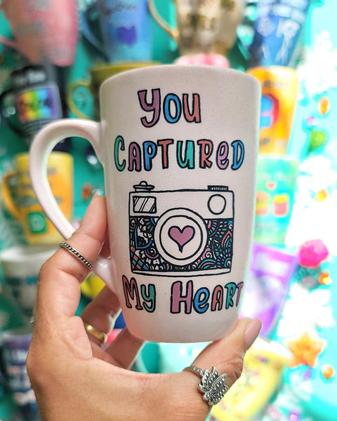 A pale pink mug with the words "you captured" and then below it a camera painted with a heart in the camera center, then below it the words "my heart"