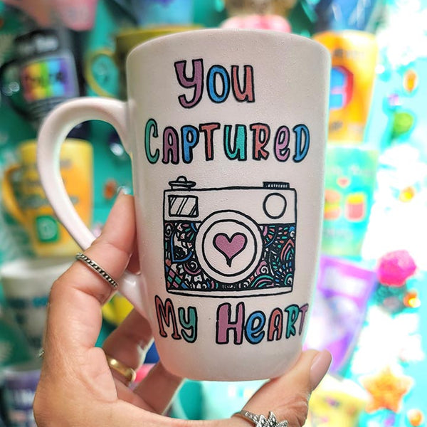 A pale pink mug with the words "you captured" and then below it a camera painted with a heart in the camera center, then below it the words "my heart"