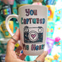 A pale pink mug with the words "you captured" and then below it a camera painted with a heart in the camera center, then below it the words "my heart"