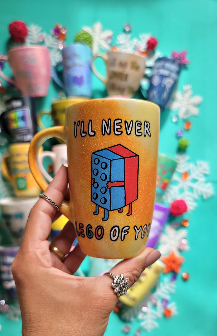 A yellow orange hand-painted mug with the words "I'll never" and below it two lego pieces hugging each other and the below it the words "LEGO of you"