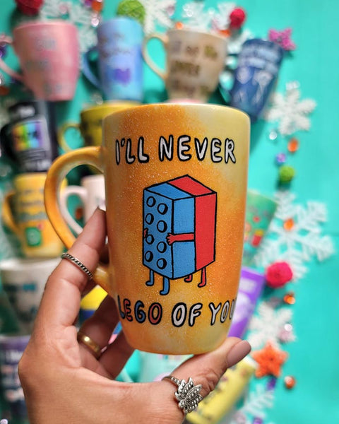 A yellow orange hand-painted mug with the words "I'll never" and below it two lego pieces hugging each other and the below it the words "LEGO of you"