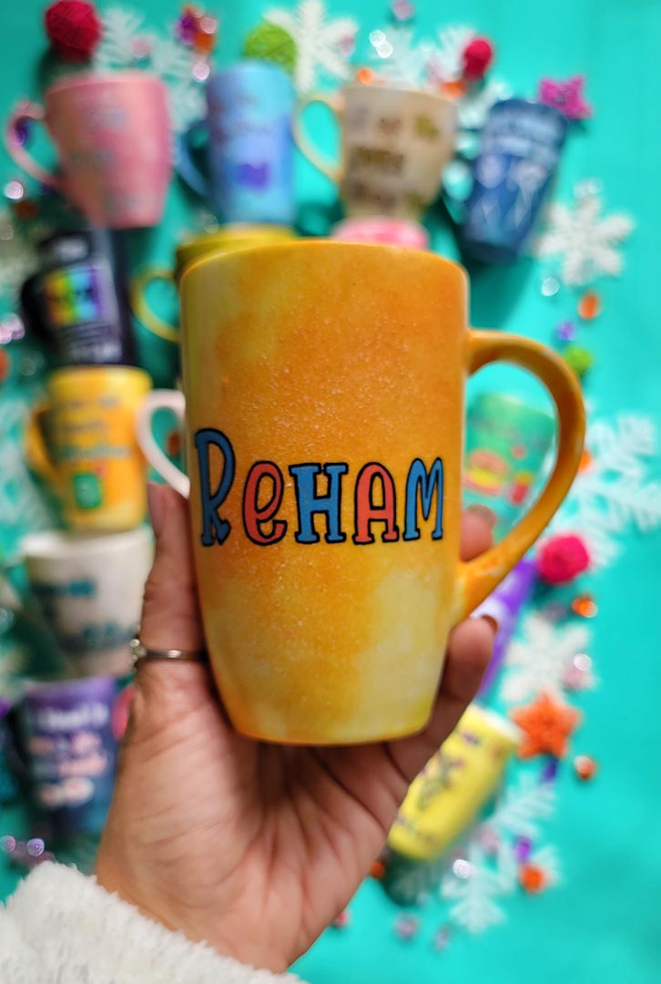 A yellow orange hand-painted mug with the name "Reham"