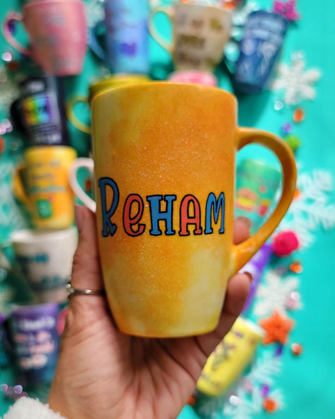 A yellow orange hand-painted mug with the name "Reham"