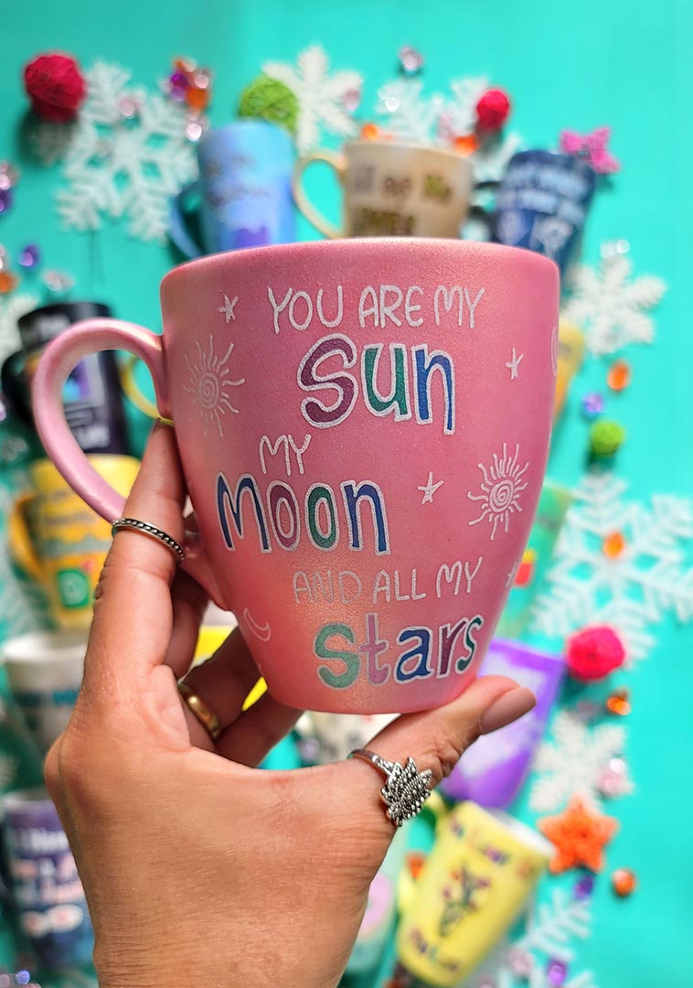 A Shimmery Pink Hand painted mug with the sentence "you are my Sun, My Moon and all my stars" with small moons stars and suns scattered all over the mug