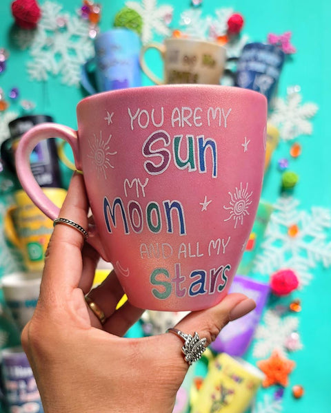 A Shimmery Pink Hand painted mug with the sentence "you are my Sun, My Moon and all my stars" with small moons stars and suns scattered all over the mug