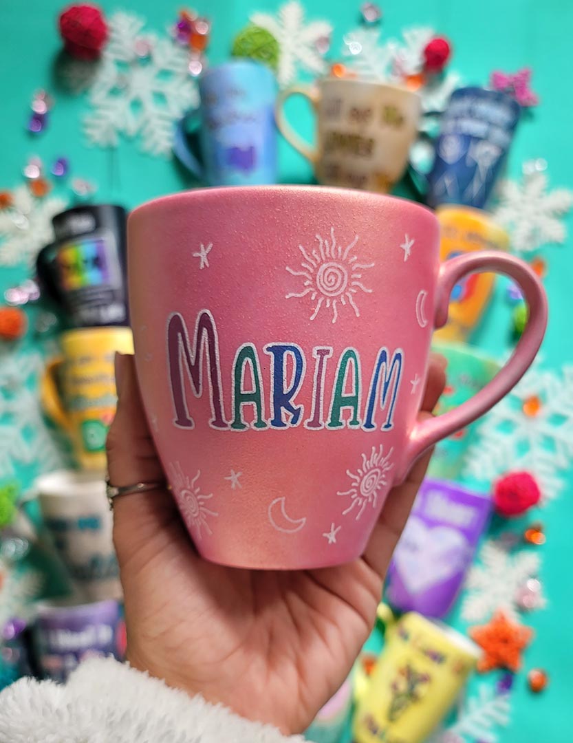 A shimmery pink hand painted mug with the name "mariam" with small moons stars and suns scattered all over the mug