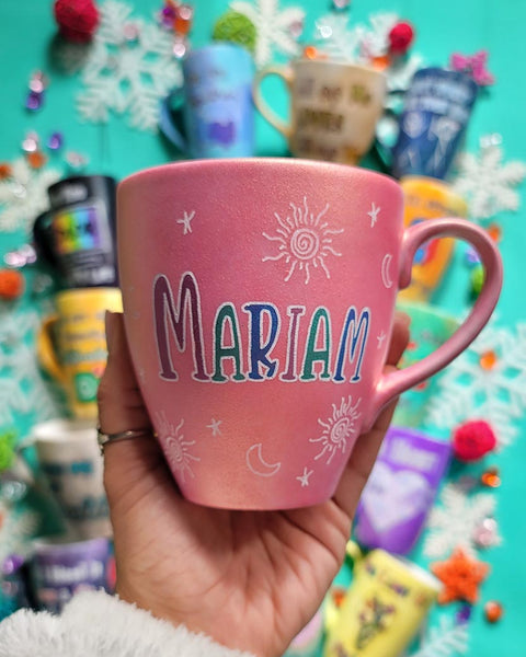 A shimmery pink hand painted mug with the name "mariam" with small moons stars and suns scattered all over the mug