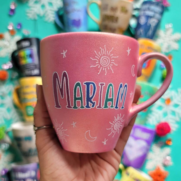 A shimmery pink hand painted mug with the name "mariam" with small moons stars and suns scattered all over the mug