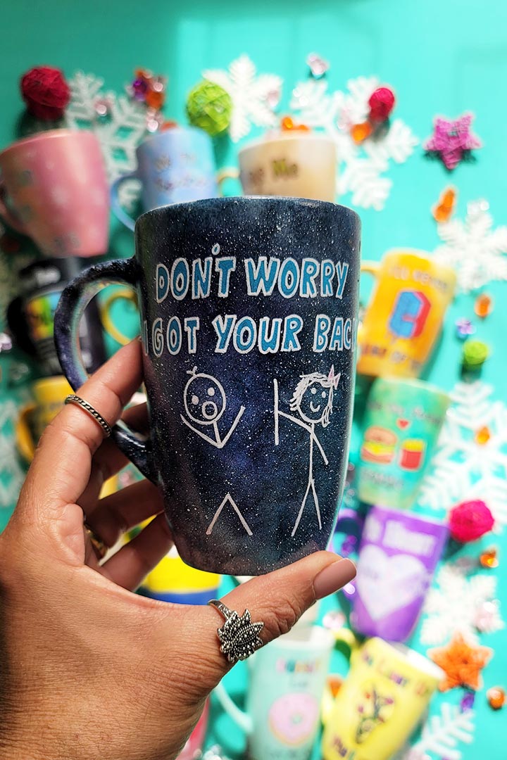 A hand painted black galaxy mug with the words "don't worry, I got your back " and a drawing of 2 stick figures with the girl holding the back of the boy