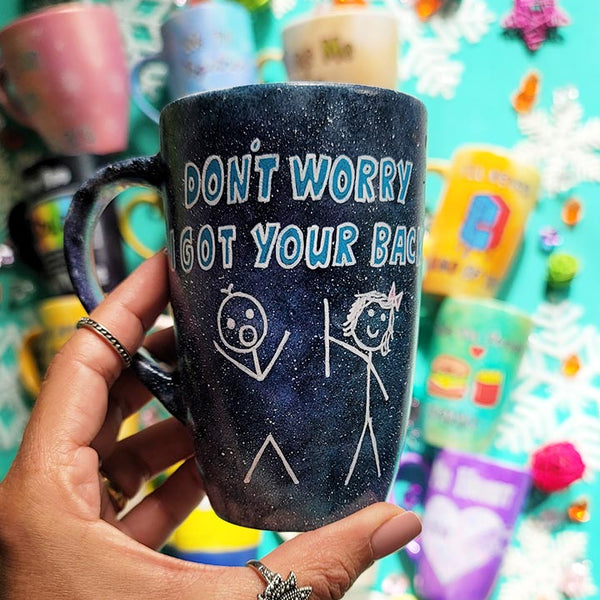 A hand painted black galaxy mug with the words "don't worry, I got your back " and a drawing of 2 stick figures with the girl holding the back of the boy