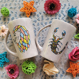 2 hand-painted taupe mugs one with the leo zodiac sign and the other with the scorpio zodiac sign