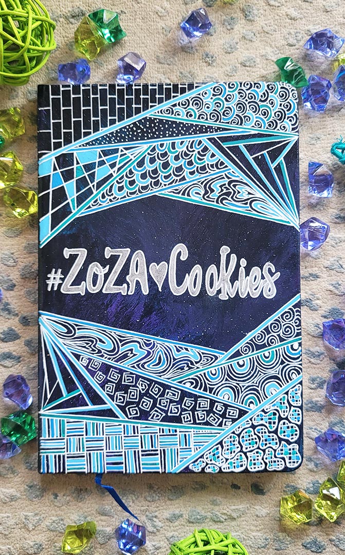 A Bluish galaxy hand painted notebook with patterns on the top and bottom and the words "Zoza heart Cookies" is written